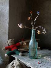 a still life arrangement featuring, on an old table, a glass bottle, withered flowers and a teddy bear slumped in a corner